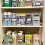 Clear plastic bins filled with various cleaning supplies organized on white shelves with handwritten labels.