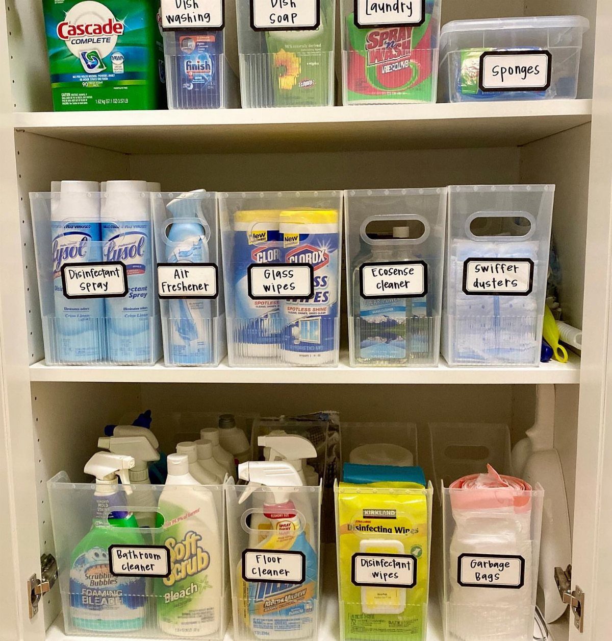 Clear plastic bins filled with various cleaning supplies organized on white shelves with handwritten labels.