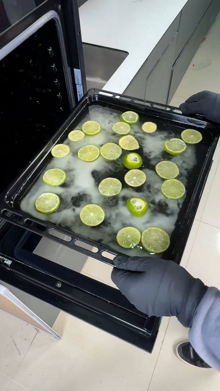 Gloves holding a black baking tray filled with white liquid and lime slices over an open oven.