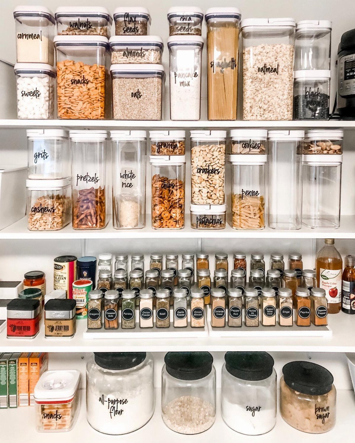 Clear plastic storage containers filled with various dry goods and spices organized on white kitchen shelves.