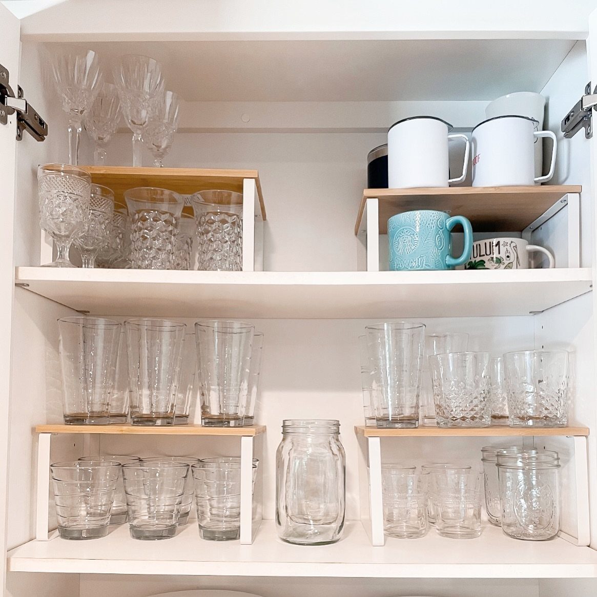 Clear glassware and mugs organized on wooden shelf risers inside white kitchen cabinets.